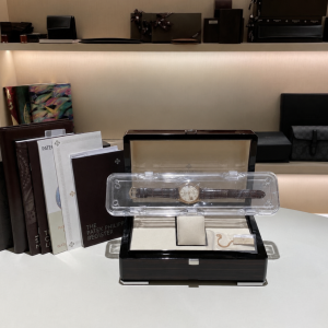 Luxurious wristwatch displayed in a clear case on a white table, with catalogs and leather boxes in the background.