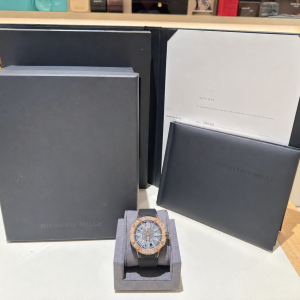 Richard Mille watch with rose‑gold bezel on a gray cushion, showcased with dark brand boxes and a certificate in the background.