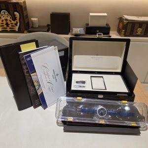 Patek Philippe watch in an open presentation box with papers and a clear plastic case on a desk in a showroom setting.