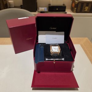 Open Cartier jewelry box with a gold-tone wristwatch, white Roman-numeral dial, and a blue strap resting in the tray beneath the watch.