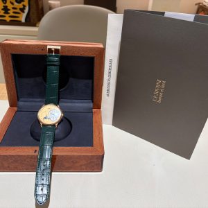 Green leather watch strap resting in an open wooden box with a dark blue interior, next to a gray document folder and a tiger artwork in the background.