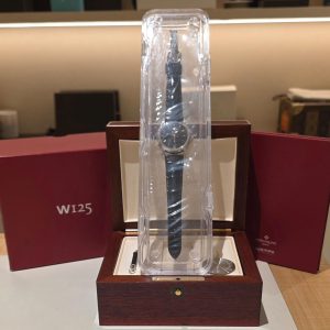 Wristwatch displayed in a tall clear acrylic case on a wooden stand with burgundy gift boxes in the background.
