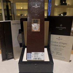 Patek Philippe watch in a tall brown presentation box on display, surrounded by certificates and packaging boxes in a shop window.