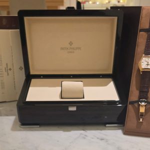 Open luxury watch box displaying a gold Patek Philippe wristwatch with a brown leather strap in a showroom display.