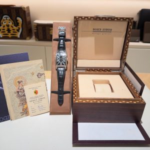 Roger Dubuis wristwatch with black leather strap in a presentation box, with certificates and booklets nearby on a table.