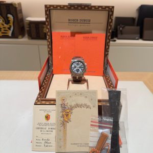Luxury wristwatch displayed in an open Roger Dubuis box with a red interior and orange booklets behind it, on a light table.