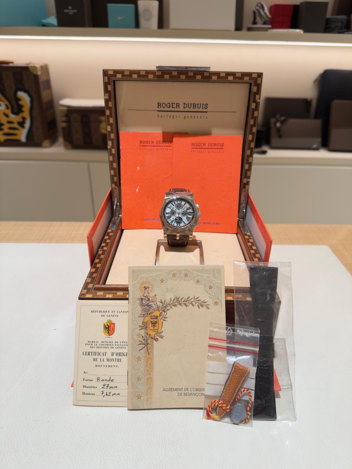 Luxury wristwatch displayed in an open Roger Dubuis box with a red interior and orange booklets behind it, on a light table.
