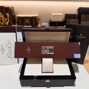 Assorted luxury watch boxes and folders on a display shelf, including a black Patek Philippe case in the foreground and branded materials in the background.