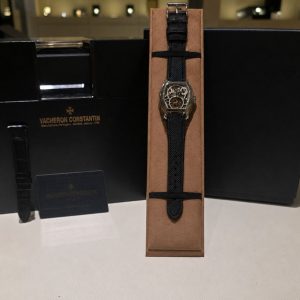 Vacheron Constantin wristwatch with a black textured strap on a brown display stand beside a black luxury box.