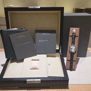 Luxury watch with brown leather strap in a brown presentation box, surrounded by black display boxes and wallets on a beige stand