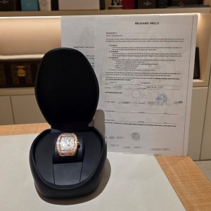 Rose-gold wristwatch in a black display case on a light desk, with a large warranty document and blurred boxes in the background