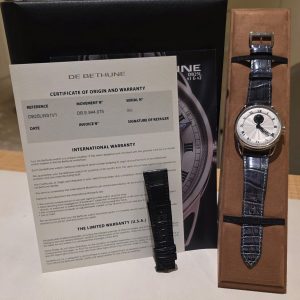 Watch with a blue crocodile strap in a brown presentation box, next to a De Bethune certificate of origin and warranty document.
