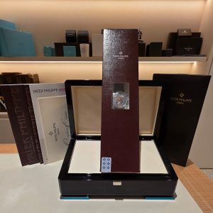 Shelf display of luxury watch boxes and brochures, including Patek Philippe branded packaging on a glossy stand and shelves behind it.