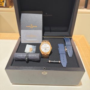 Open black presentation box displaying a gold Vacheron Constantin wristwatch with blue strap options, warranty cards, and accessories arranged on a velvet tray.