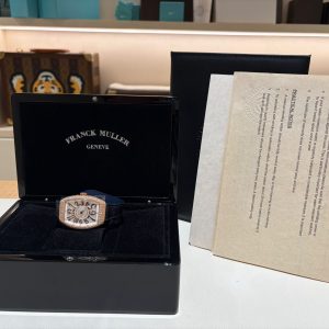 Open black display box holding a rose-gold Franck Muller Genève wristwatch, with a beige document partially visible to the right.