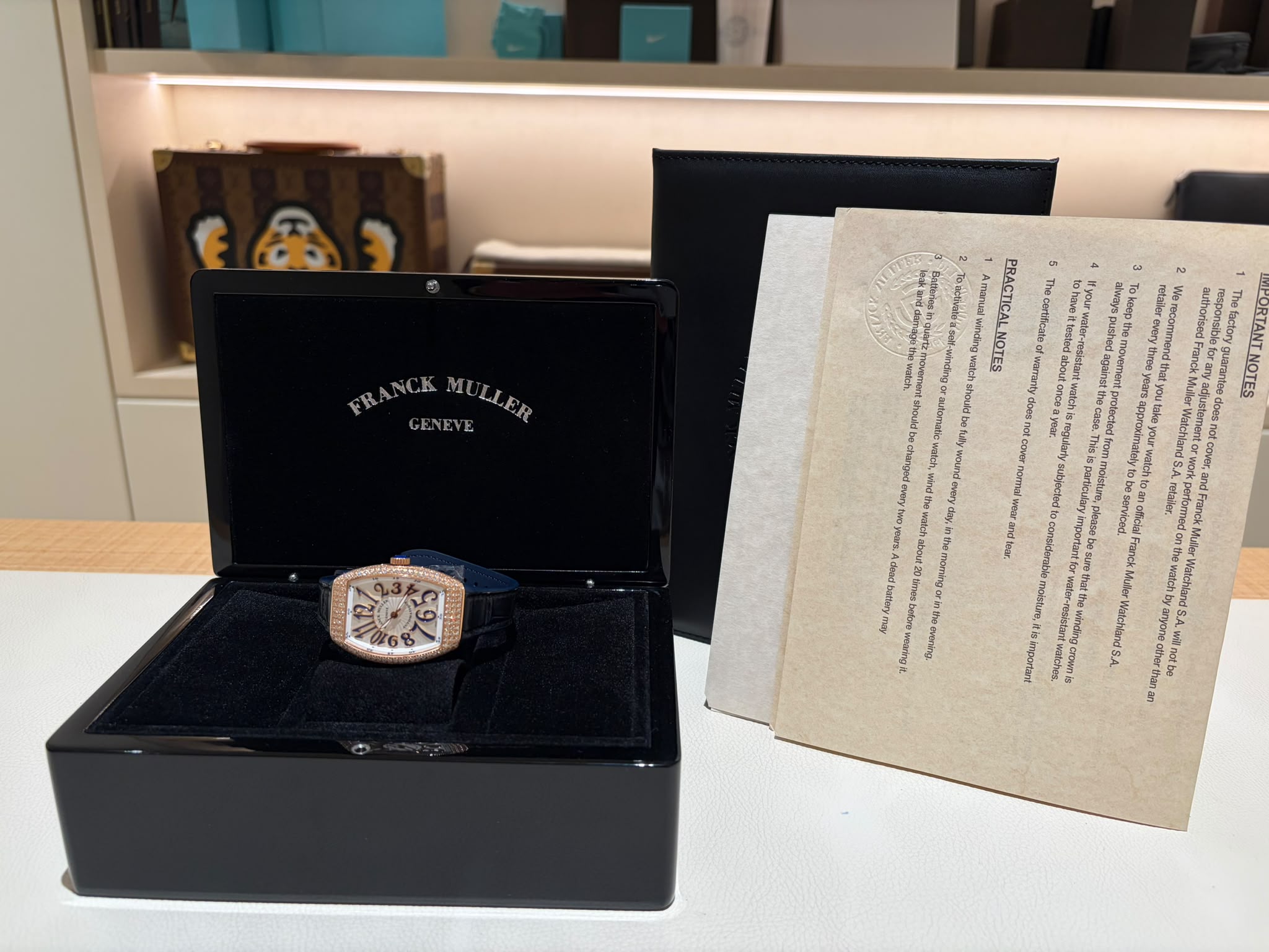 Open black display box holding a rose-gold Franck Muller Genève wristwatch, with a beige document partially visible to the right.