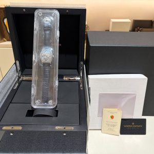 Luxury wristwatch in clear plastic display case inside an open black presentation box with a textured exterior and protective padding nearby, and certificate cards visible nearby.