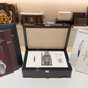 Open black presentation box showing a stainless steel Patek Philippe wristwatch on a white cushion, with brochures and catalogs nearby.