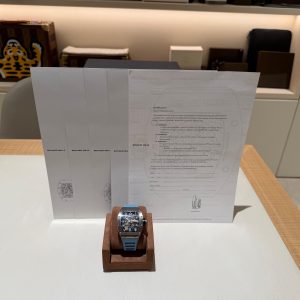 Watch on brown display stand with a textured gray catalog behind; showroom shelf with a tiger-patterned case in the background