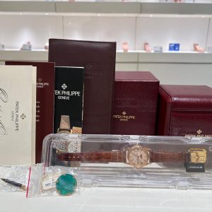 Assorted Patek Philippe watch boxes in burgundy and cream on a display table, with a brown leather pouch and a clear plastic case showing a gold-tone wristwatch inside.