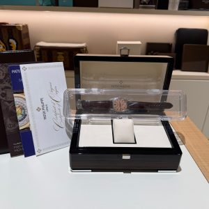 Patek Philippe watch in a clear display case on a white counter, surrounded by brochures and a leather folder in a showroom setting