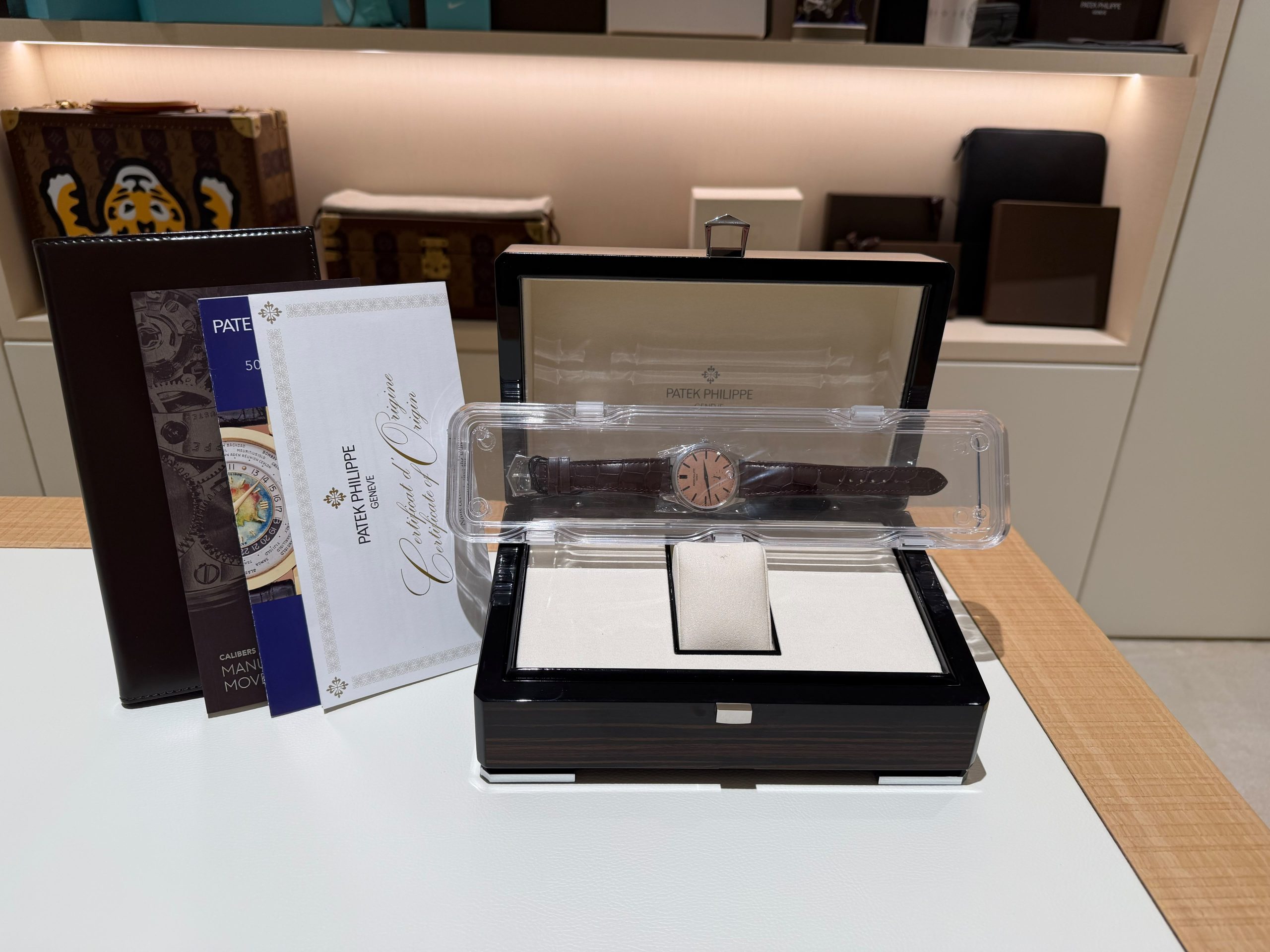 Patek Philippe watch in a clear display case on a white counter, surrounded by brochures and a leather folder in a showroom setting