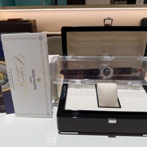 Open black watch display case with a round-faced Patek Philippe watch on a white cushion; brochures stand beside it, on a showroom counter