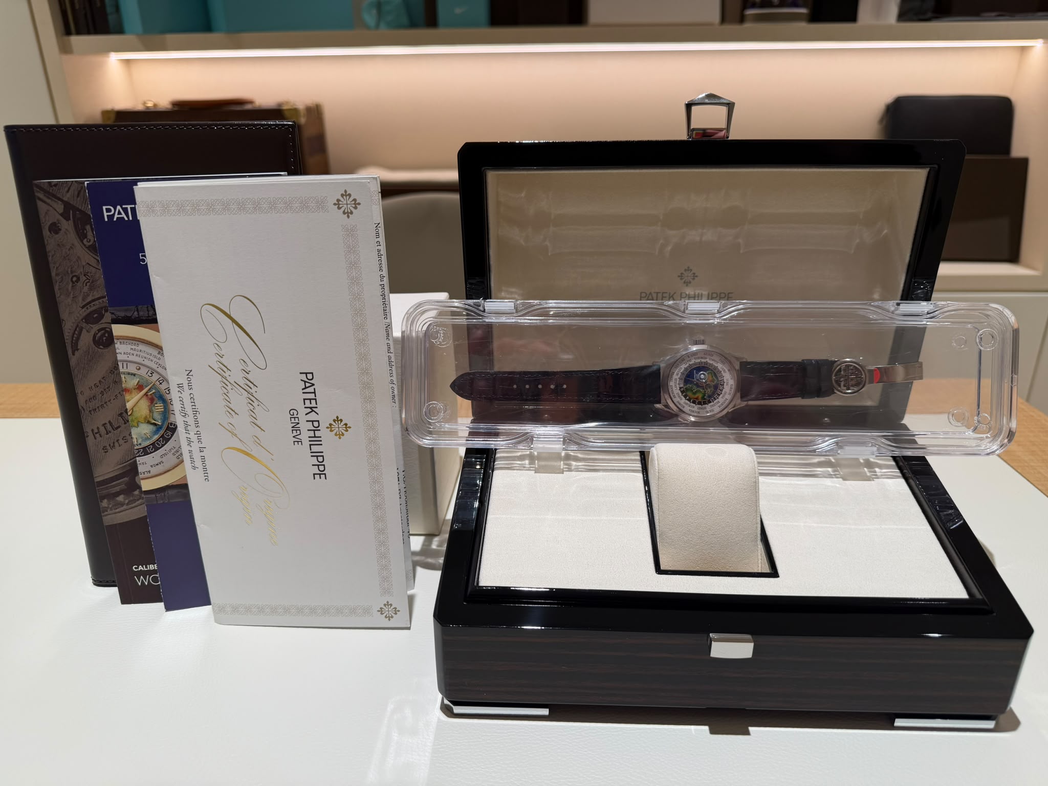Open black watch display case with a round-faced Patek Philippe watch on a white cushion; brochures stand beside it, on a showroom counter