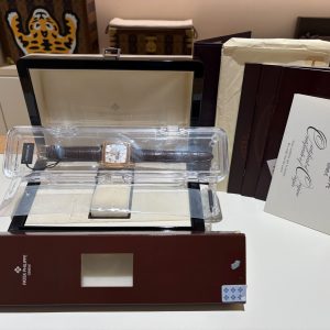 Patek Philippe wristwatch in a clear plastic display case on a dark wooden box, with brochures nearby in a shop setting.