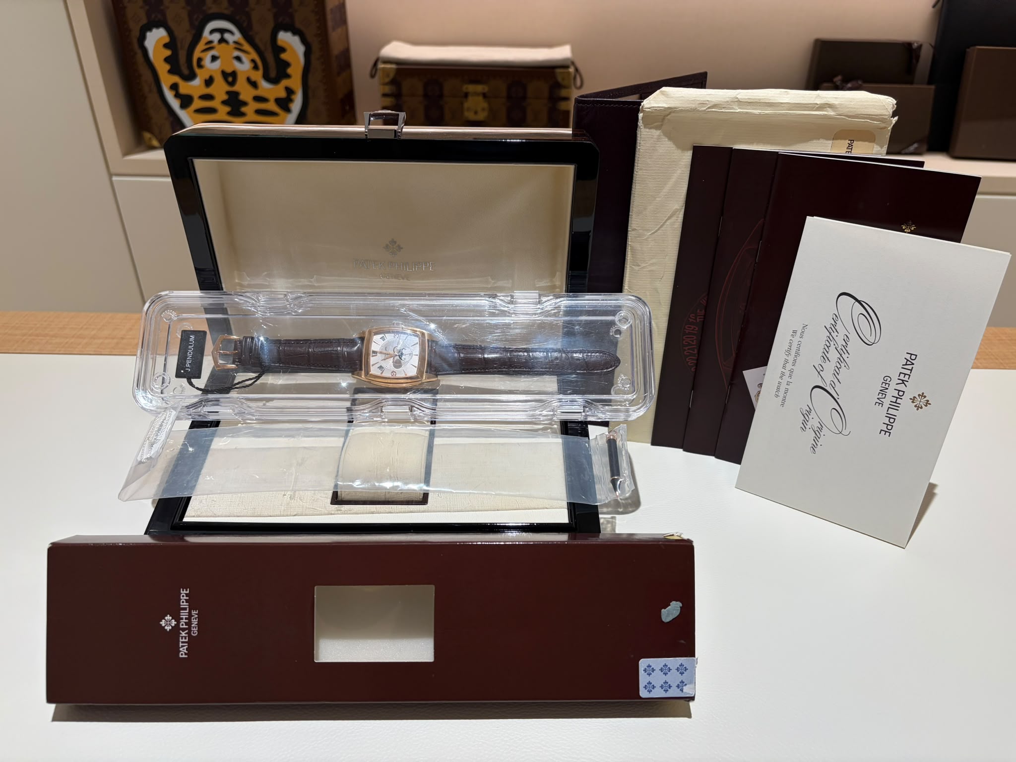 Patek Philippe wristwatch in a clear plastic display case on a dark wooden box, with brochures nearby in a shop setting.
