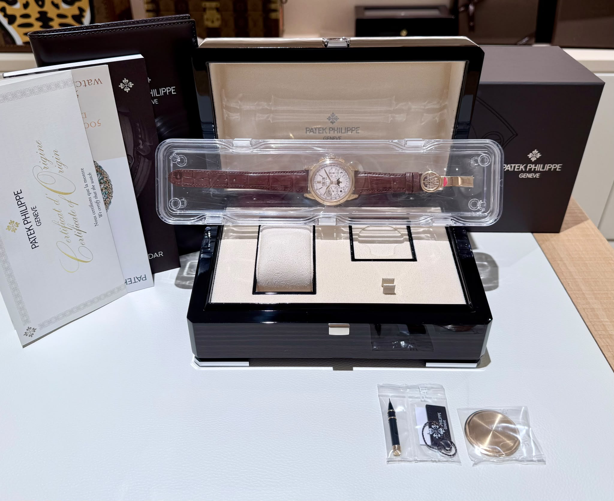 Patek Philippe watch with brown leather strap in a clear plastic display case, surrounded by black presentation box and papers.