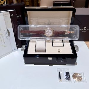 Patek Philippe wristwatch with brown leather strap in a clear plastic display case on a black presentation box, surrounded by manuals.