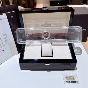 Patek Philippe wristwatch with a brown leather strap displayed in an open black presentation box with a clear plastic protective case around it.