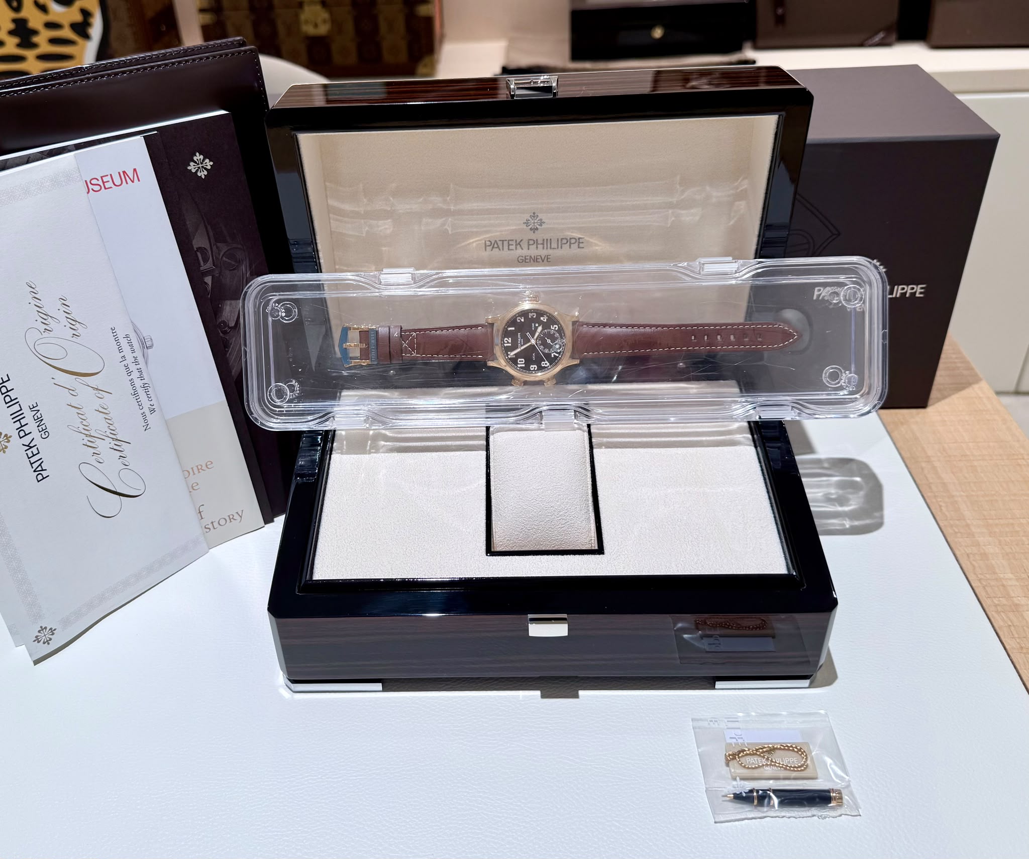 Patek Philippe wristwatch with a brown leather strap displayed in an open black presentation box with a clear plastic protective case around it.