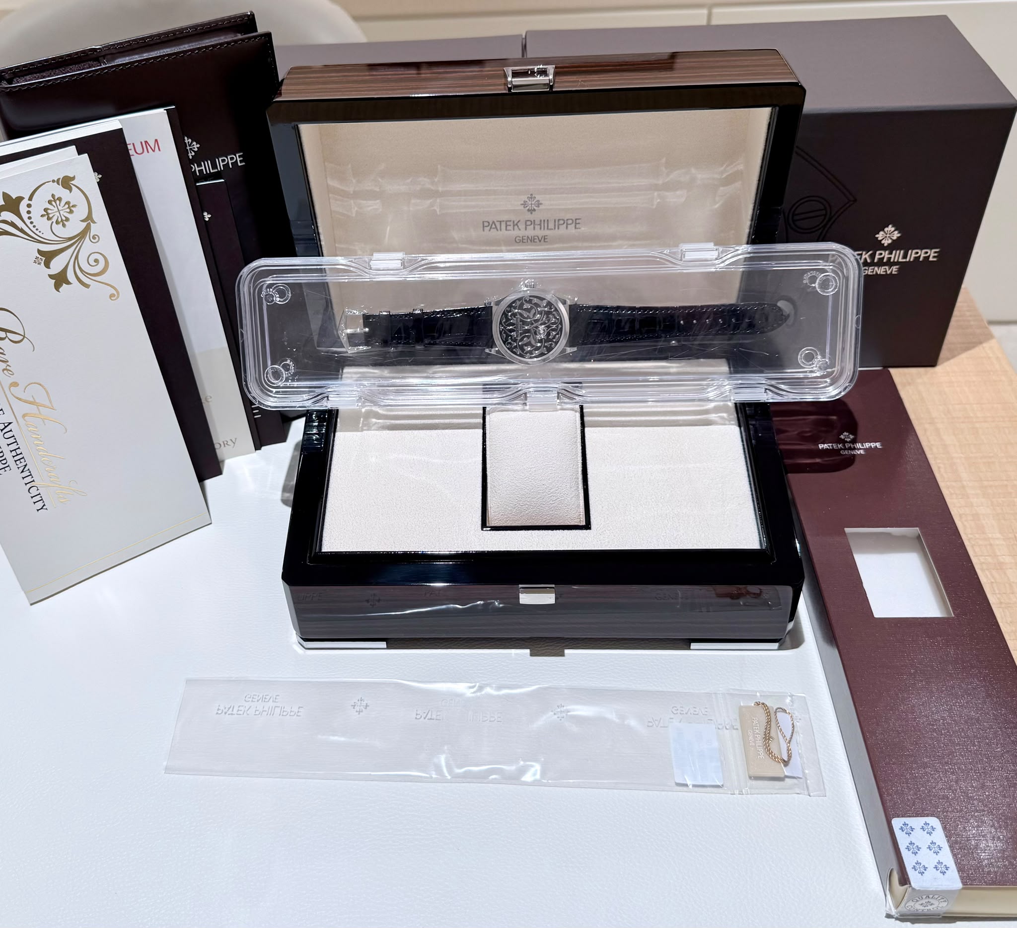 Patek Philippe watch in opened display box with clear plastic case and accompanying burgundy packaging boxes.