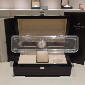 Patek Philippe wristwatch in a glass display case at a jewelry store, with black and beige presentation boxes behind it.