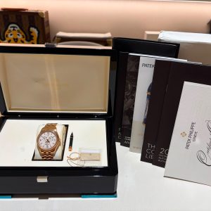 Rose-gold wristwatch in an open black presentation box with white interior, alongside brand brochures and catalogs on the right.
